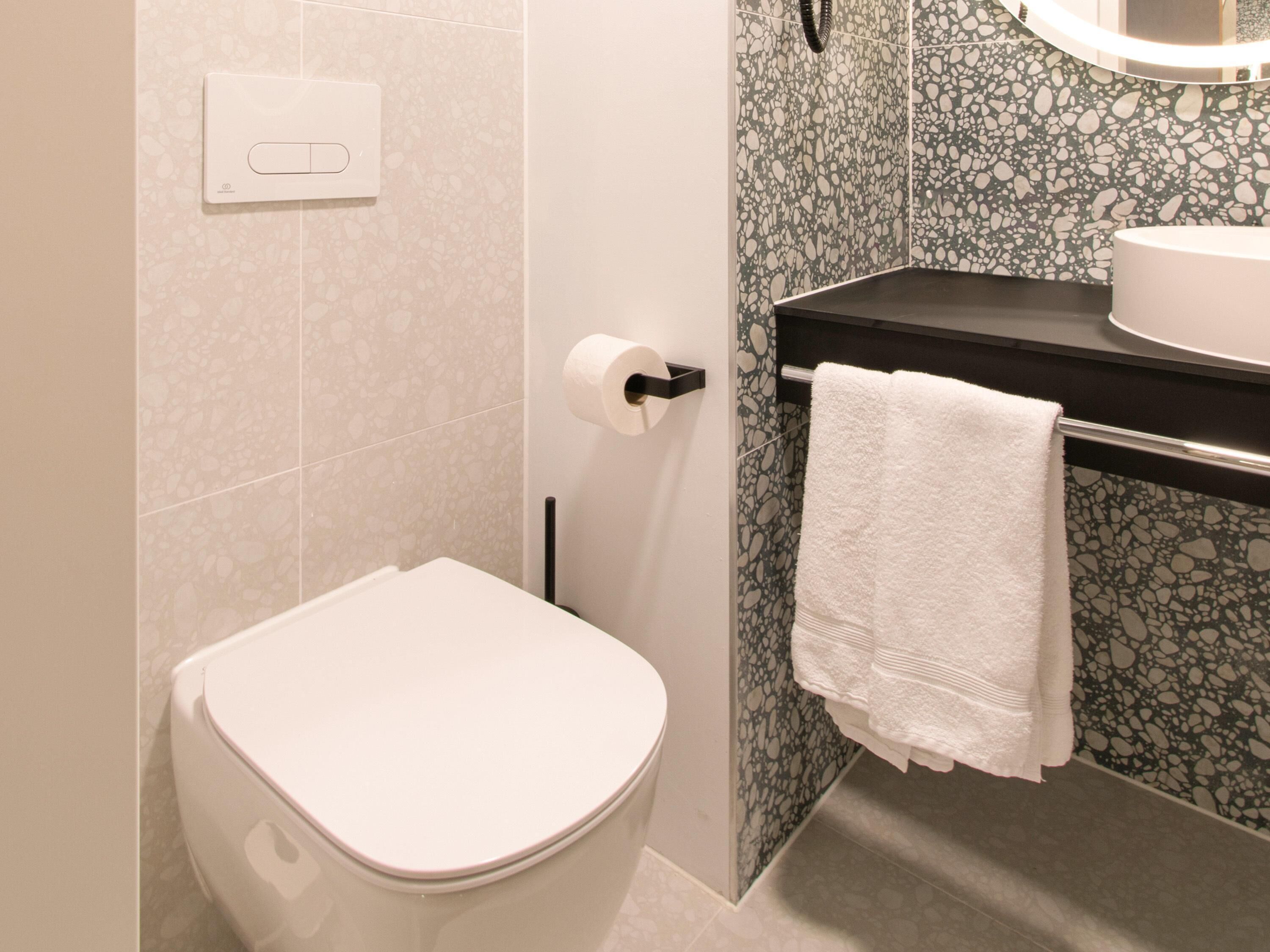 Un baño con inodoro, toallero con toalla blanca, dispensador de papel higiénico y lavamanos con espejo arriba.