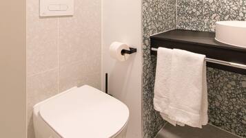 Un baño con inodoro, toallero con toalla blanca, dispensador de papel higiénico y lavamanos con espejo arriba.