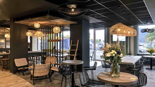 A modern cafe interior with wicker furniture, round tables, and hanging wicker lamps. - Ibis Styles Bredene  (Bredene)