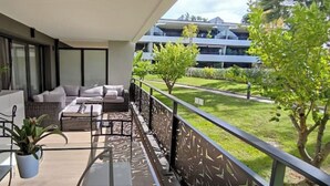 Un balcon avec un canapé, une chaise et un pot de fleurs. On y trouve une balustrade, ainsi qu’une vue sur une pelouse verdoyante et des arbres.