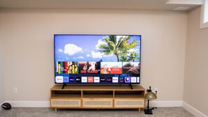 Un televisor de pantalla plana sobre un soporte de madera con un estante debajo, mostrando una interfaz de plataforma de transmisión con varias miniaturas de programas.