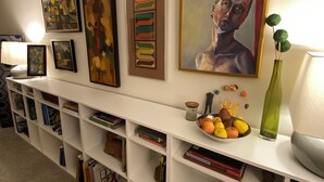 A room with a white wall adorned with various framed artworks, a white bookshelf filled with books and decorative items, and a small table with a bowl of fruit and a vase. - Trilith Studios Area Lakefront  (Sharpsburg)