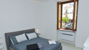 A modern bedroom with a grey bed, white nightstands, a window with a view of the street, and a radiator. - Angel's house in Sorrento coast (Meta)