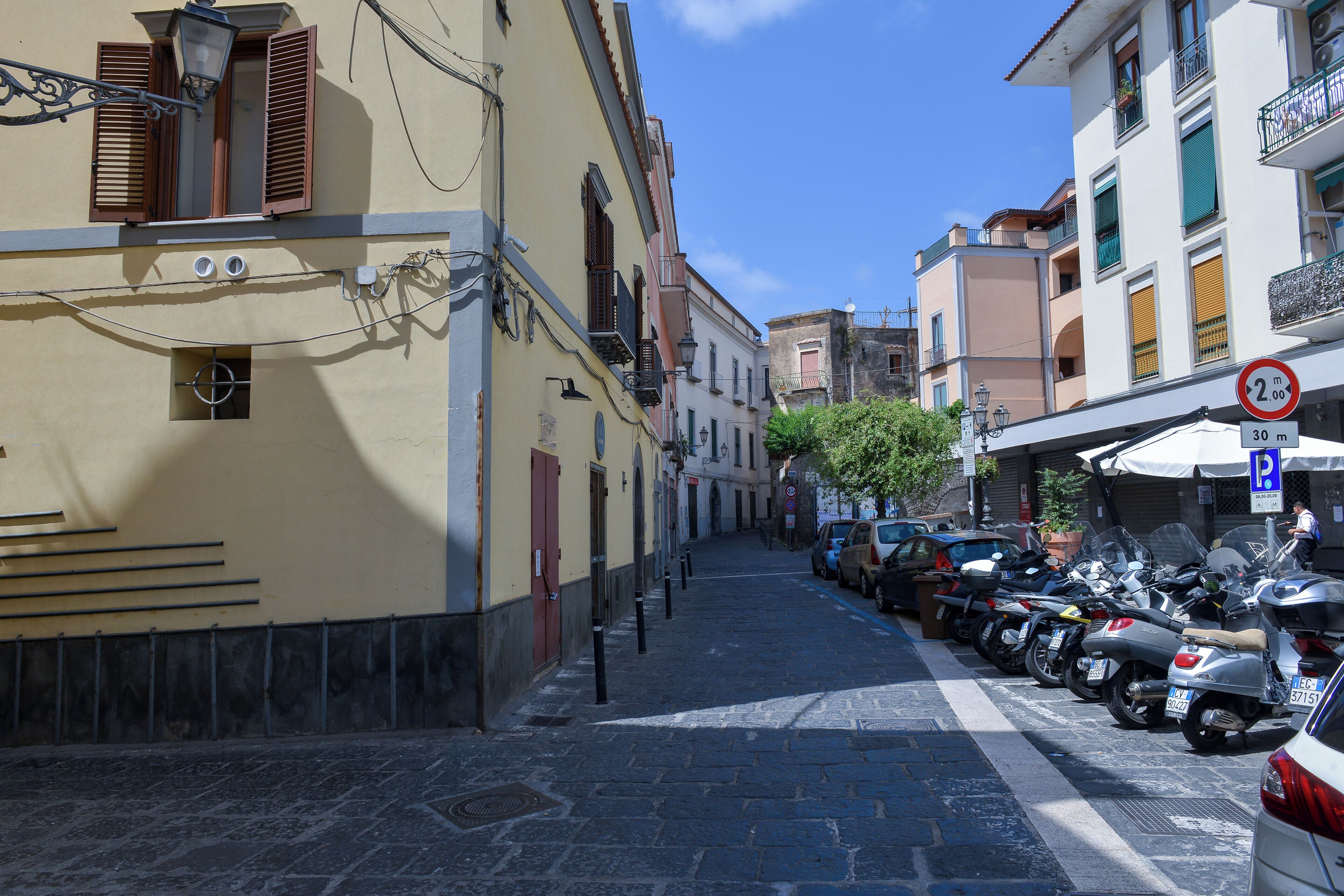 A narrow street lined with parked motorcycles and cars, with buildings featuring balconies and shutters.