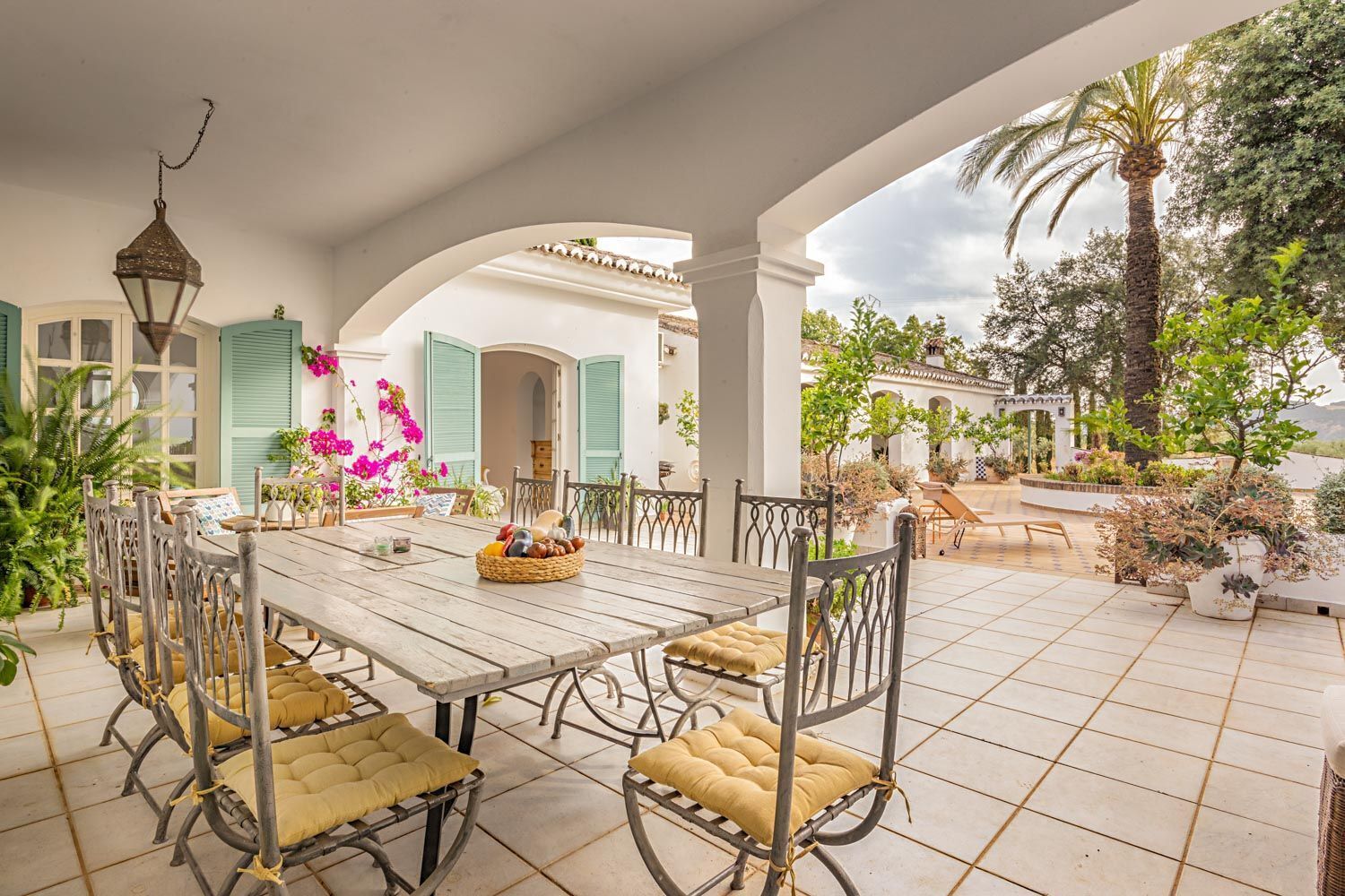 An outdoor patio with a wooden table and metal chairs, surrounded by greenery and a view of a garden.