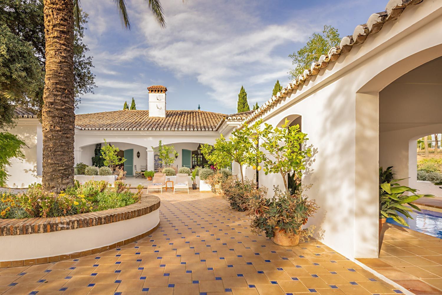 A courtyard with a tiled floor, white walls, and a tiled roof. There are potted plants and a bench.