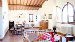 A spacious living room with a high wooden ceiling, a dining area with a wooden table and chairs, and a cozy seating area with a plaid sofa and a vase of flowers. - Podere Belvedere - Superbe et Grande Villa de Campagne Avec Piscine Privée (Carmignano)
