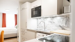 A modern kitchen with a built-in microwave, oven, and sink. There is a bed with orange curtains and a wooden chair visible in the background. - House of Blooms (Fulda)