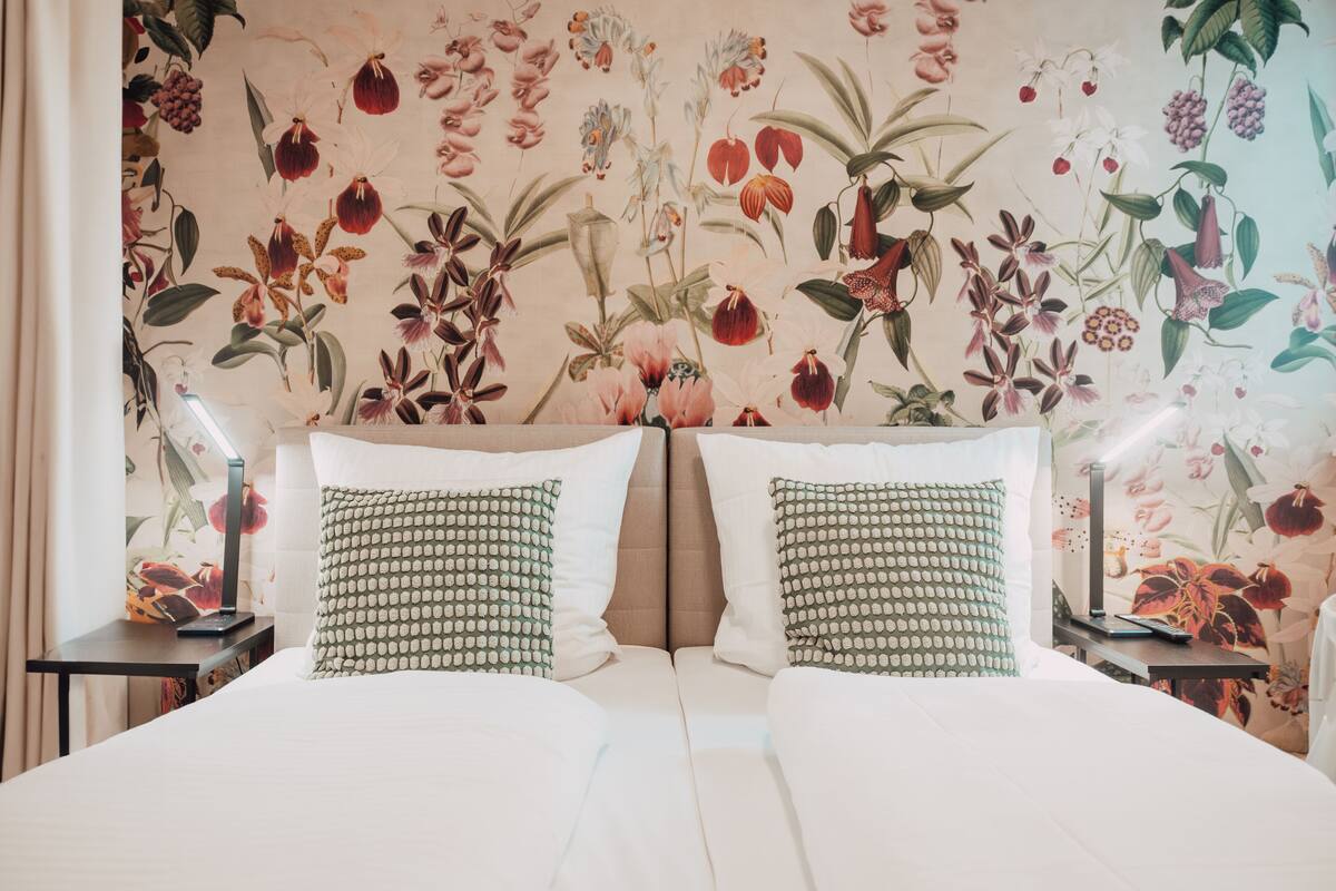 A double bed with two pillows, a nightstand with a lamp, and a floral wallpaper.