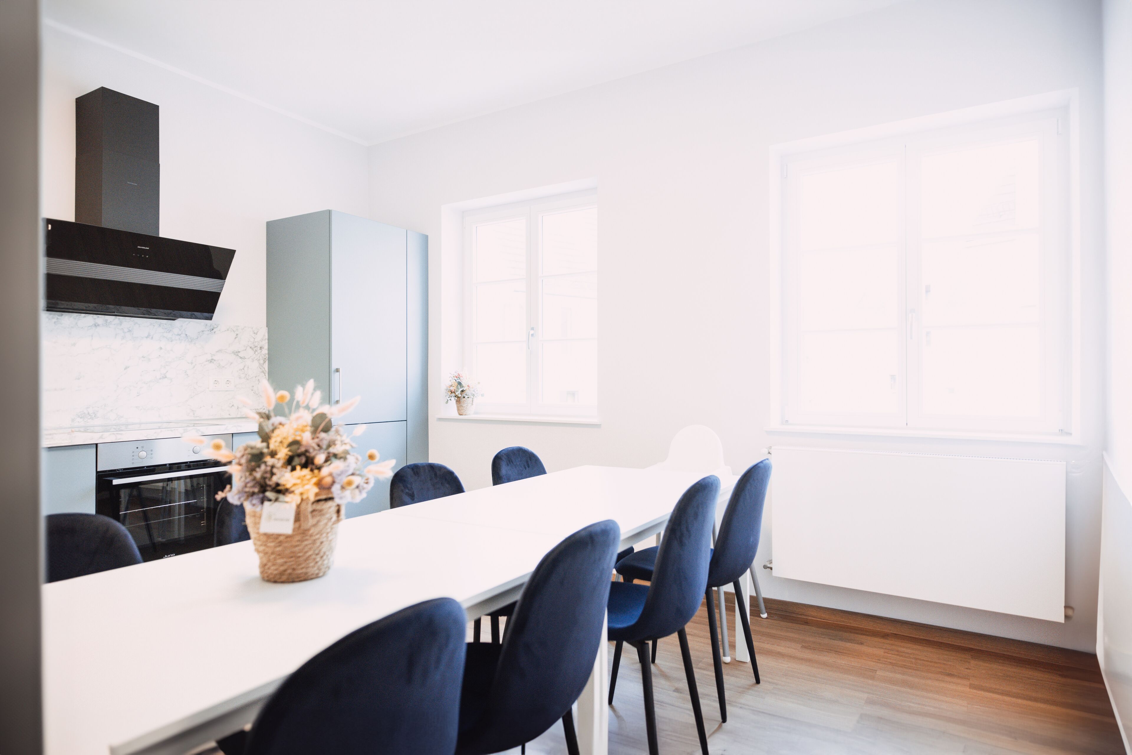 Eine moderne Küche mit weißer Arbeitsplatte, einem eingebauten Backofen und einer schwarzen Dunstabzugshaube. Ein Esstisch mit blauen Stühlen und eine Blumenvase stehen bereit.