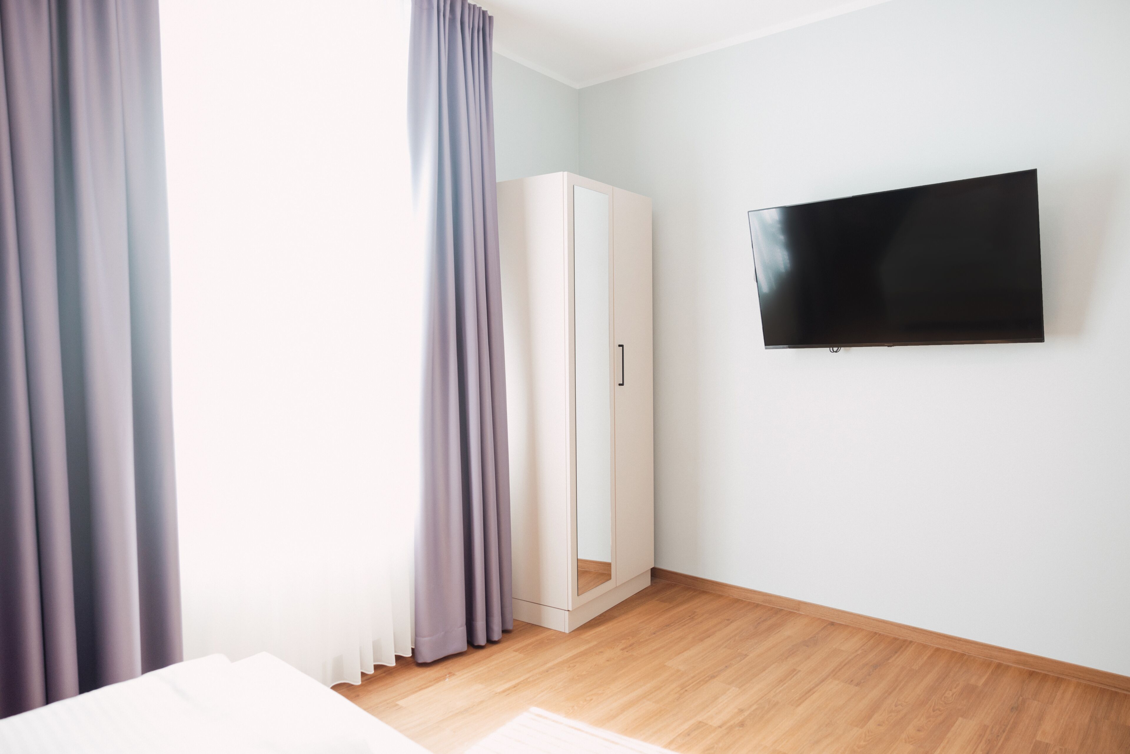 Una habitación con cama, armario, televisor en la pared y ventana con cortinas.