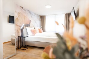 A modern hotel room with a large bed, a flat-screen TV mounted on the wall, a bedside table with a lamp, and a view of the city through large windows. - House of Blooms (Fulda)