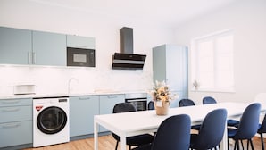 A modern kitchen with light blue cabinets, a white countertop, a built-in microwave, a washing machine, and a dining area with a white table and dark blue chairs. - House of Blooms (Fulda)