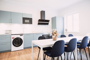 Cozinha moderna com armários azul claro, bancada branca, micro-ondas embutido, máquina de lavar e um espaço de refeições com mesa branca e cadeiras azul escuro.