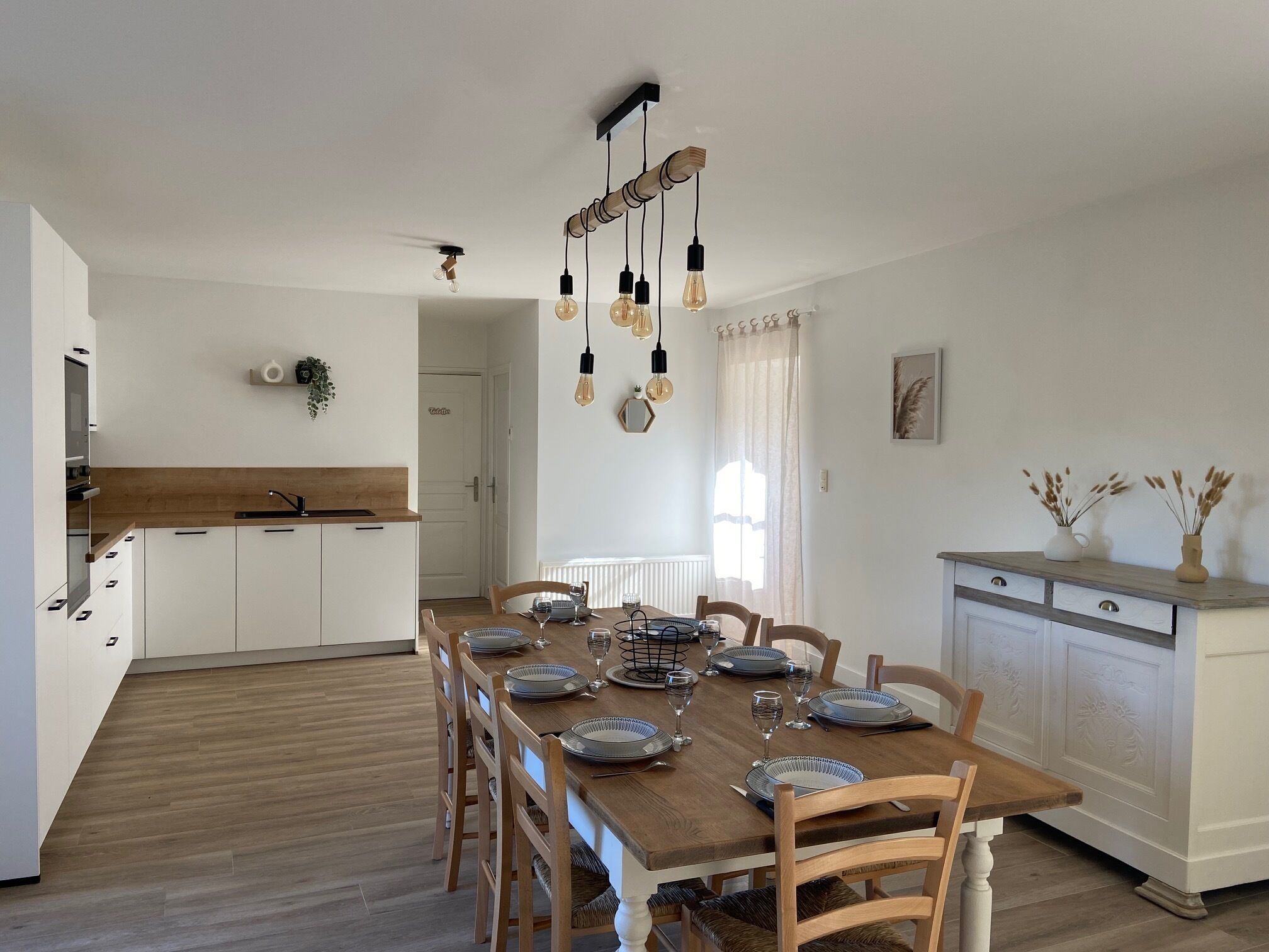 Una cocina moderna con armarios blancos, una mesa de comedor de madera para cuatro personas y una lámpara de techo con focos expuestos.