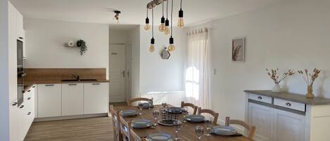 Una cocina moderna con armarios blancos, una mesa de comedor de madera para cuatro personas y una lámpara de techo con focos expuestos.