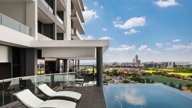 A modern hotel with a rooftop infinity pool, a glass balustrade, and a view of the cityscape and park. - Illovo Central 1023 (Sandton)