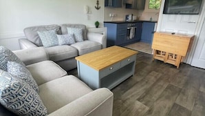 A living room with a grey sofa, a wooden coffee table, and a kitchen area with blue cabinets and a TV mounted above the door. - Villa 19 Lanteglos 2 bed sleep 5 (Lanteglos)