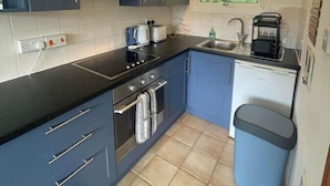 A compact kitchen with grey cabinets, a black countertop, a built-in oven, a small dishwasher, and a window with a view of greenery. - Villa 19 Lanteglos 2 bed sleep 5 (Lanteglos)