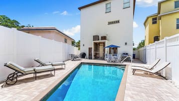 A rectangular swimming pool with lounge chairs and a table on one side, surrounded by a white fence.