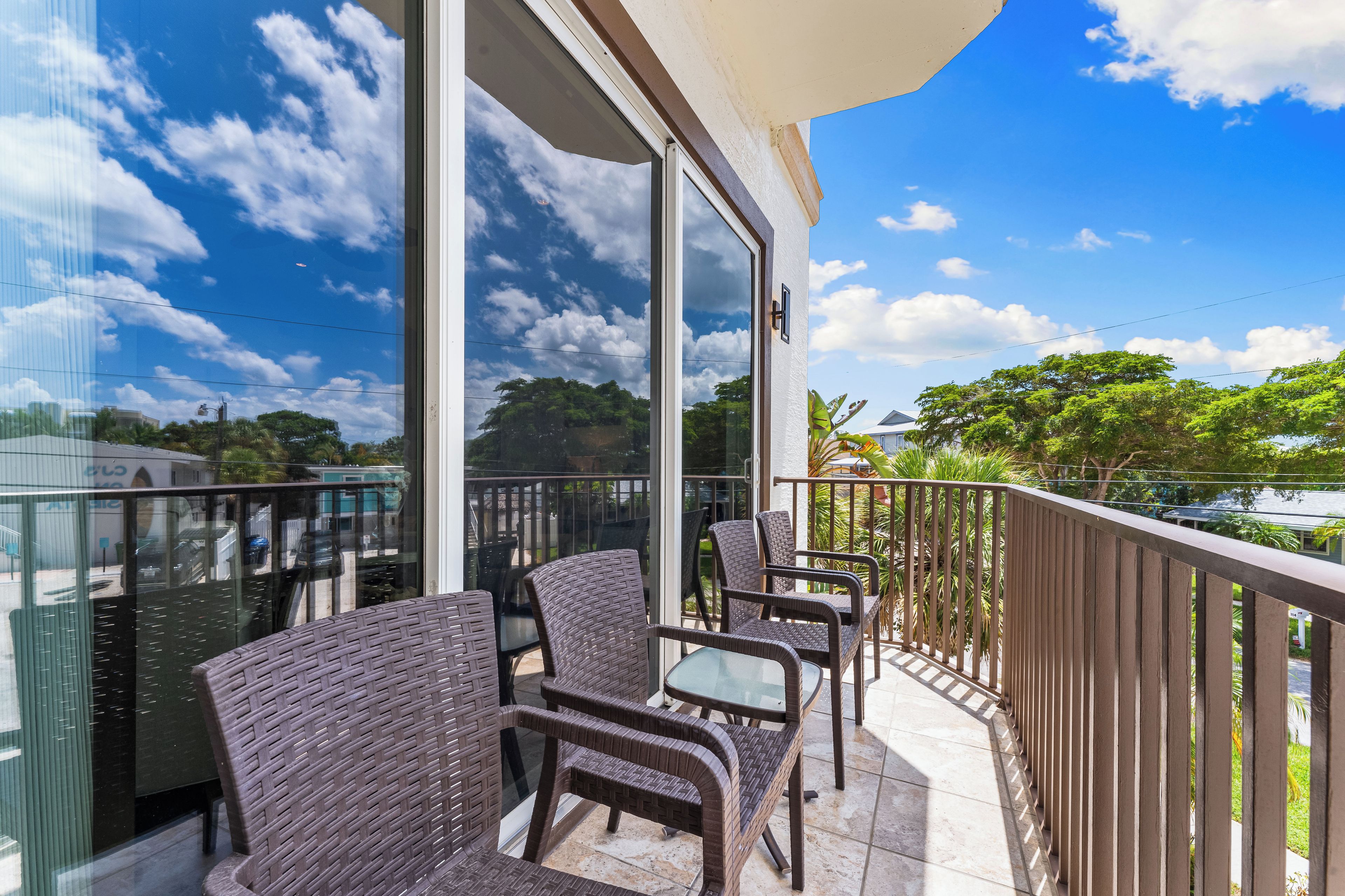 A balcony with wicker chairs and a glass table, overlooking a residential area with trees and houses.