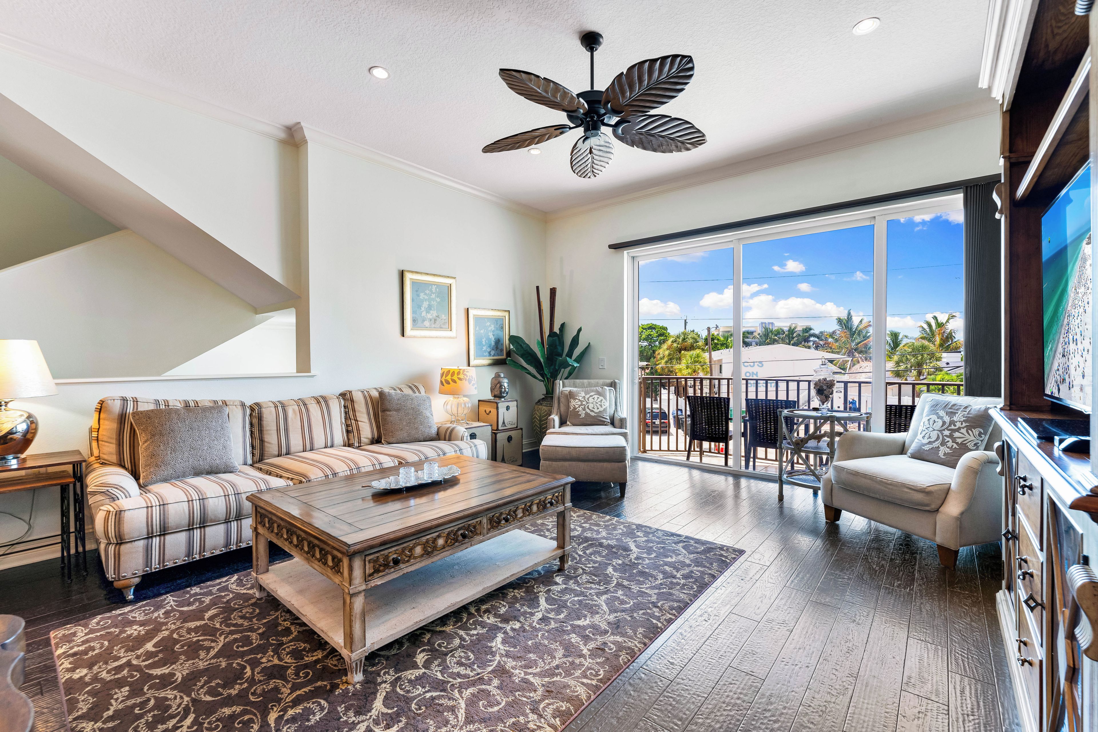 A spacious living room with a large sofa, a coffee table, and a ceiling fan. There is a sliding glass door leading to an outdoor area with a view of palm trees.