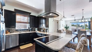 A modern kitchen with dark cabinetry, a central island, and a dining area with patterned upholstery. - Beachside of the Village by Beachside (Siesta Key)