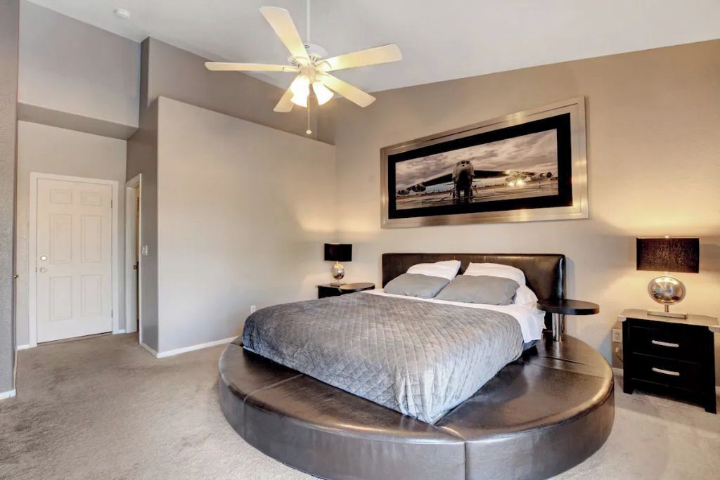 Quarto com cama curva, ventilador de teto, quadro na parede e mesa de cabeceira com luminária.