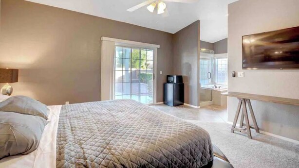 Une chambre à coucher comprenant un lit, un ventilateur de plafond, un téléviseur et une porte-fenêtre coulissante donnant sur un balcon.