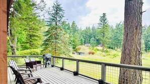A wooden deck with outdoor seating and a grill, overlooking a forested area with a large tree and houses in the background. - Lookout @ SeaAcres -Beach Access (San Juan County)