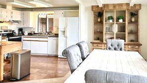 A modern kitchen with white cabinets, a dining area with a wooden table and grey upholstered chairs, and a built-in wooden shelving unit with plants. - Lookout @ SeaAcres -Beach Access (San Juan County)