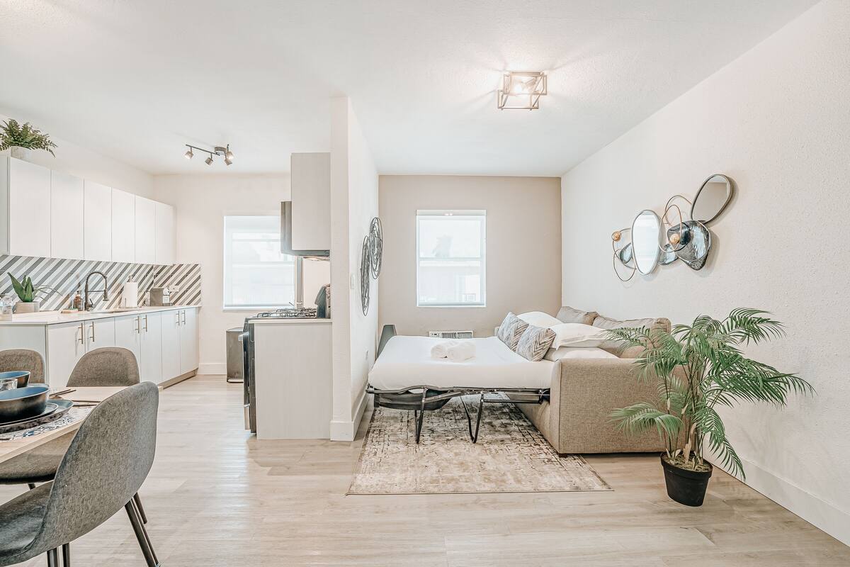 A modern apartment with a kitchen, dining area, and a bedroom with a bed and a potted plant.