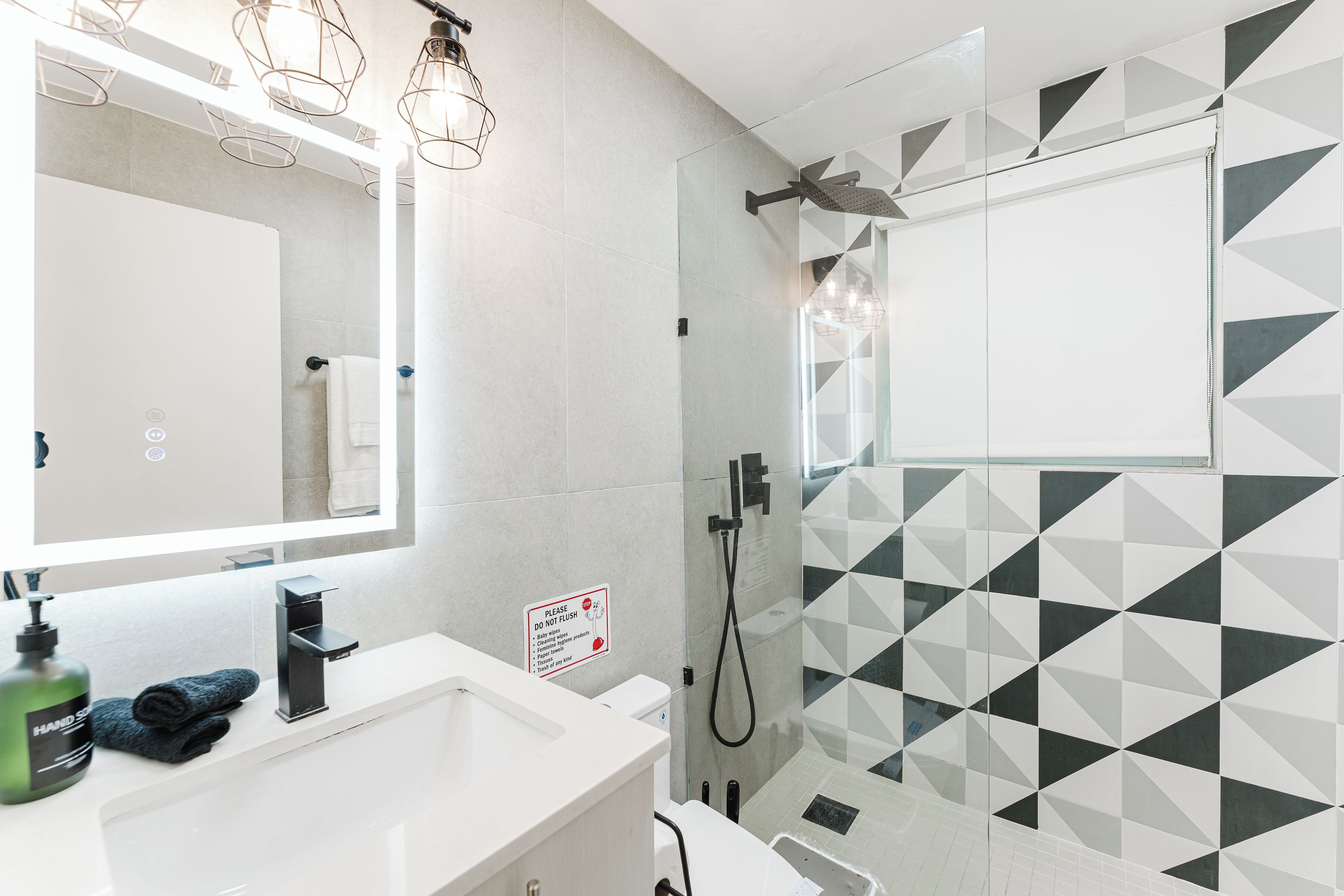 Ein modernes Badezimmer mit einem weißen Waschbecken, einem Spiegel, einer Dusche mit geometrischem Fliesendesign und einem Schild mit der Aufschrift: Nicht spülen.