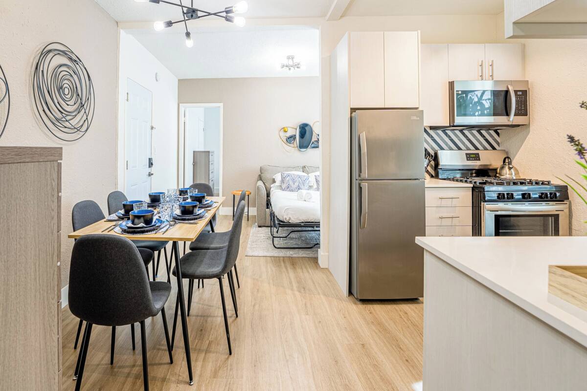 A modern kitchen with stainless steel appliances, a dining area with a wooden table and chairs, and a living area with a sofa and a TV.