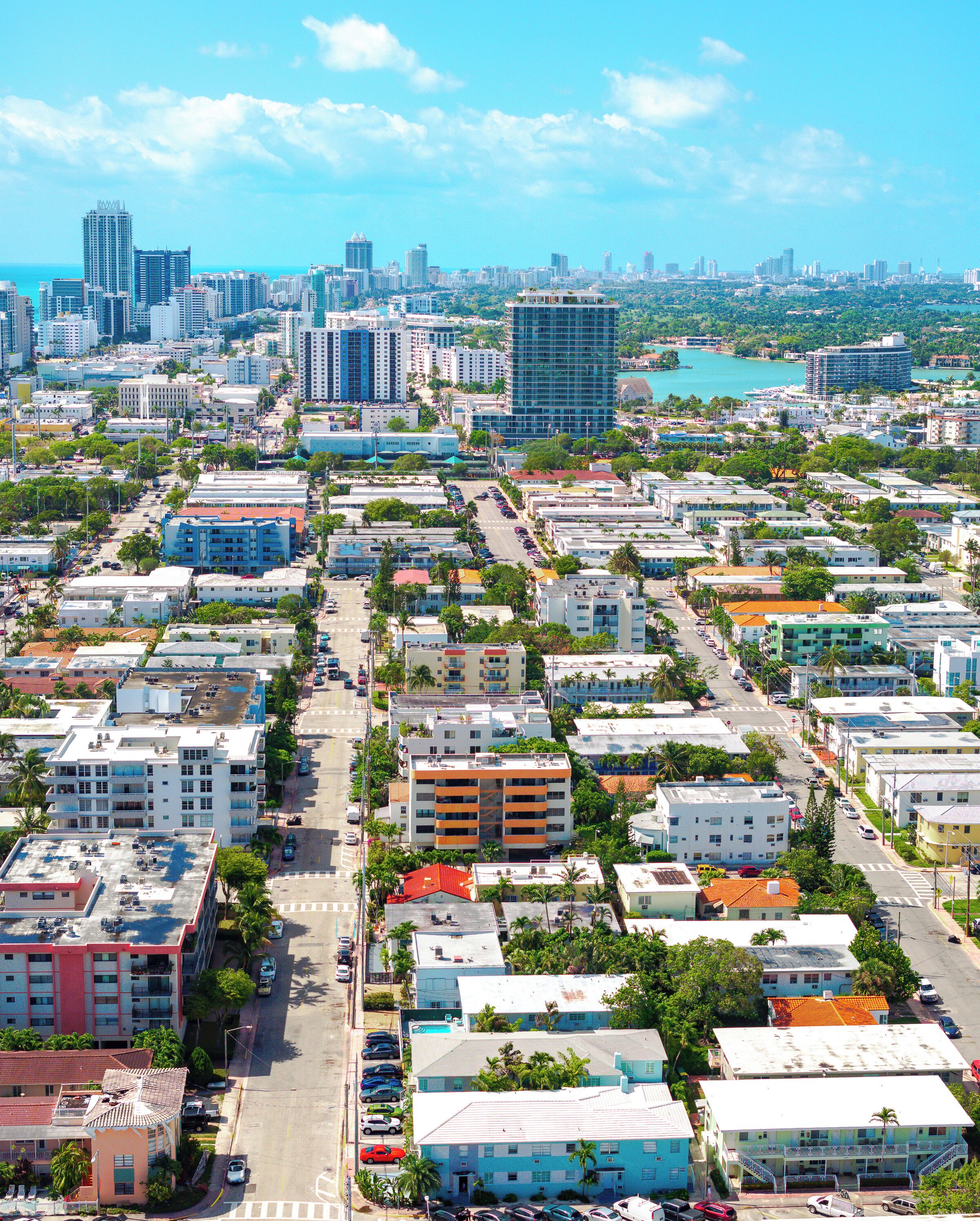 An aerial view of a cityscape with a mix of high-rise buildings and smaller structures, a clear blue sky, and a body of water in the distance.