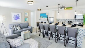 A modern kitchen with a central island, bar stools, and a dining area with a grey sofa and striped ottoman. - PRAIA Hotel Boutique Miami Beach (Miami Beach)
