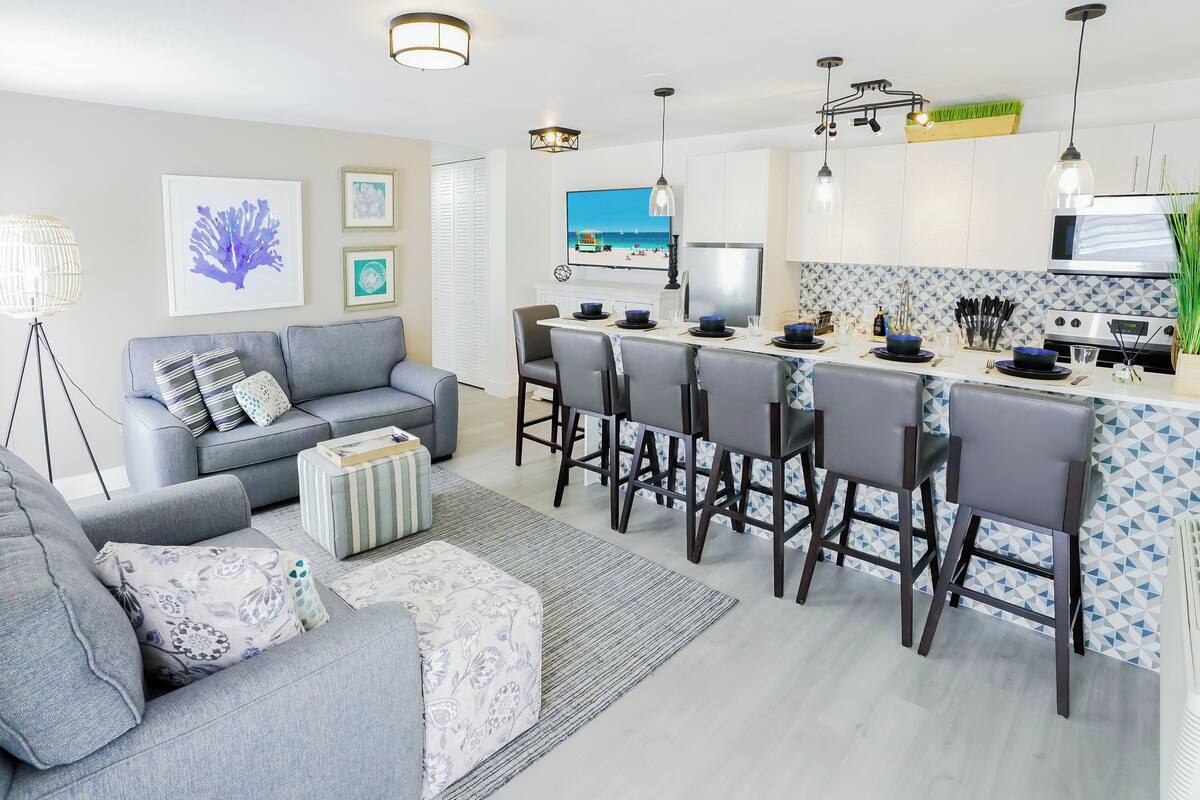 A modern kitchen with a central island, bar stools, and a dining area with a grey sofa and striped ottoman.