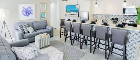 A modern kitchen with a central island, bar stools, and a dining area with a grey sofa and striped ottoman.