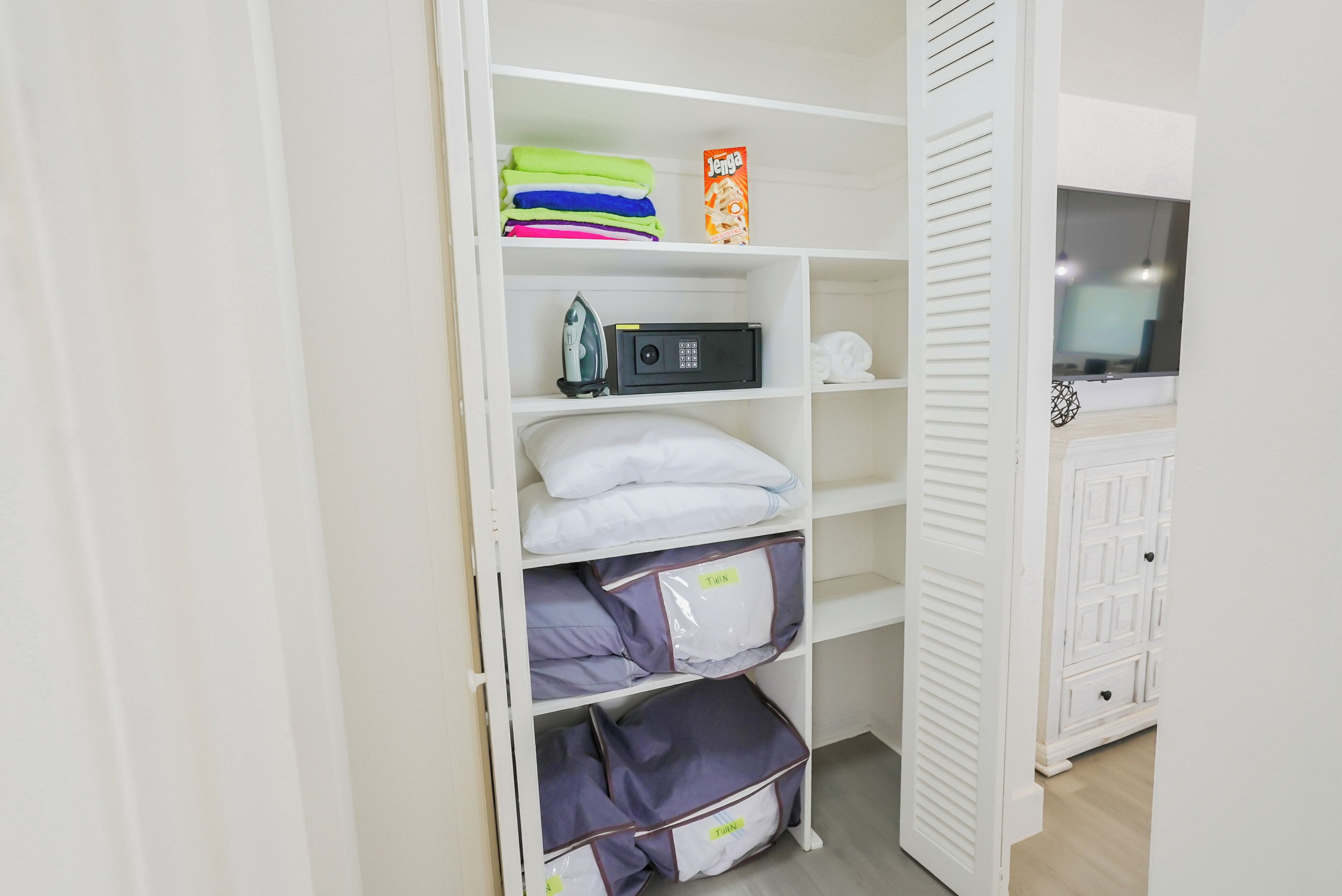 Ein weißer Kleiderschrank mit Regalen, der gefaltete Kleidung, eine Waschmittelschachtel, ein Bügeleisen und eine Mikrowelle enthält.