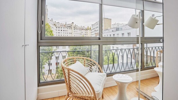 A balcony with a wicker chair and a small table, overlooking a cityscape with buildings and a clear sky. - Degas Appart 1 - Two Bedroom Apartment, Sleeps 6 (Paris)
