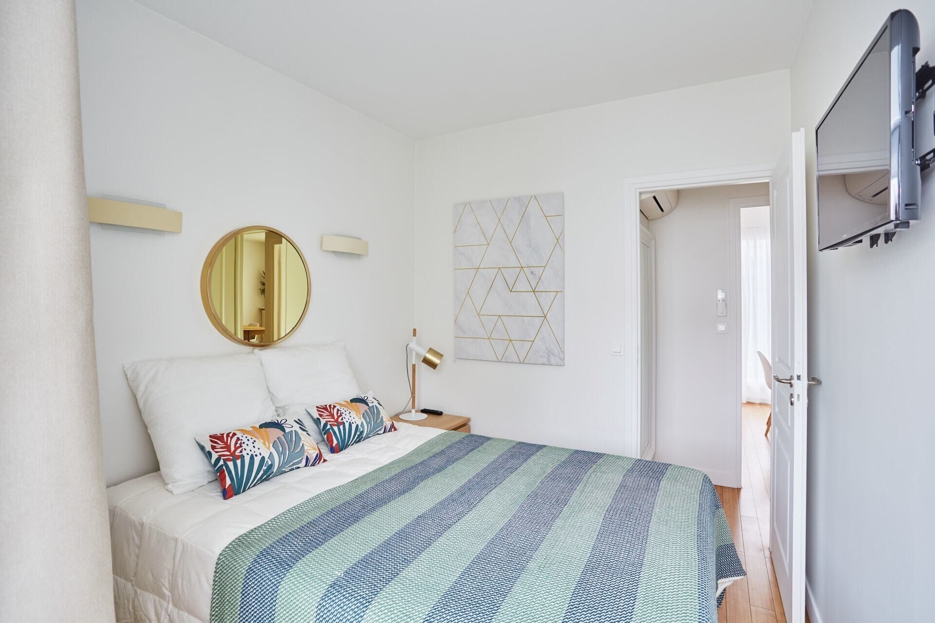 A bedroom with a bed, a round mirror, a geometric wall art, and a door leading to another room.