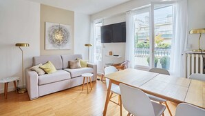 A modern living room with a sofa, a dining table, and chairs. There is a TV mounted on the wall, a floor lamp, and a painting on the wall. - Degas Appart 1 - Two Bedroom Apartment, Sleeps 6 (Paris)