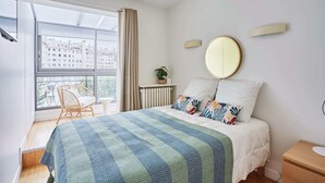 A bedroom with a bed, a window with curtains, a chair, a bedside table, and a wall-mounted mirror. - Degas Appart 1 - Two Bedroom Apartment, Sleeps 6 (Paris)