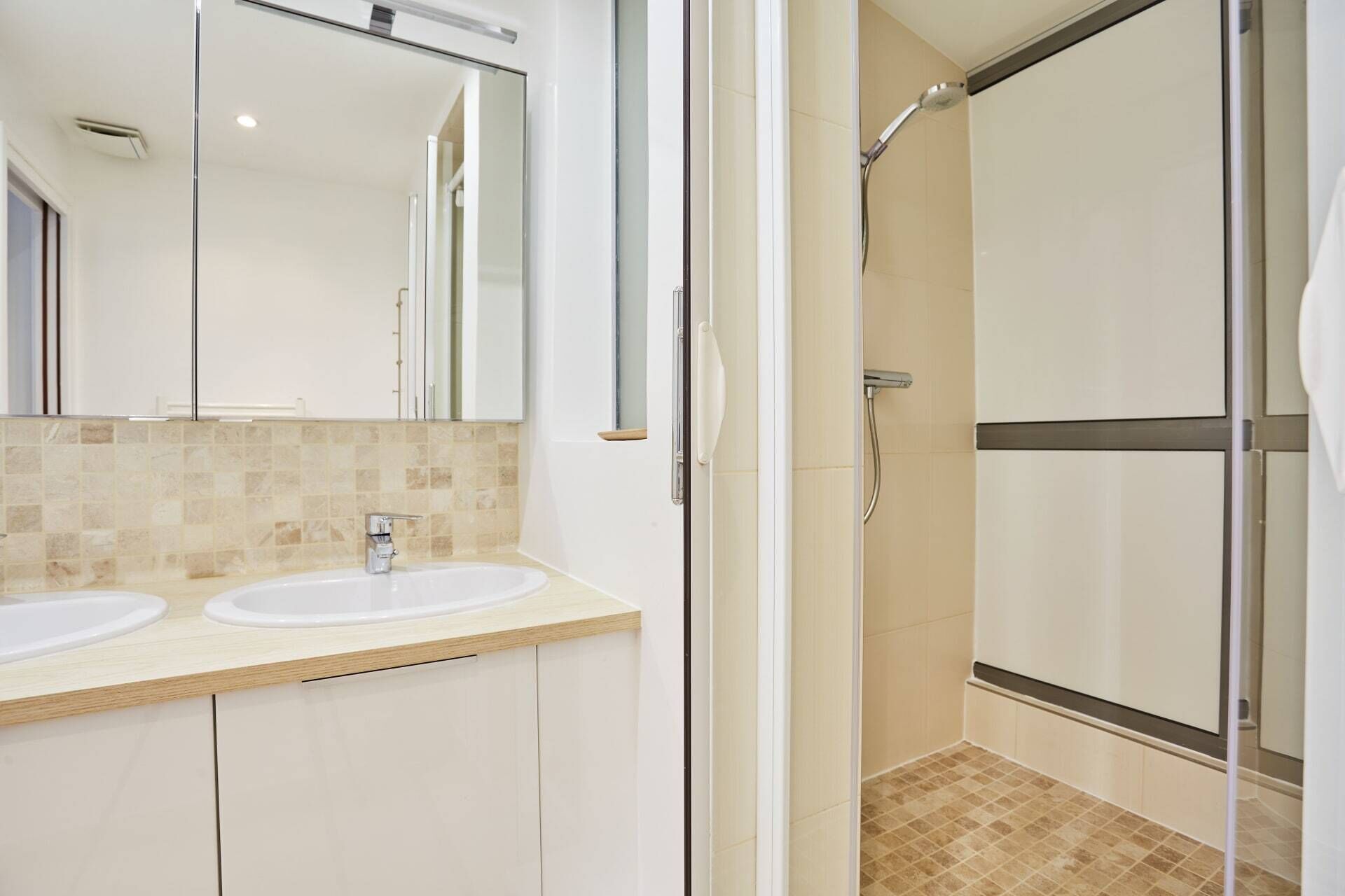 Un baño con dos lavamanos, un espejo y zona de ducha con mampara de vidrio.