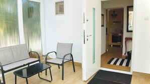 Ein Zimmer mit einer Couch, zwei Stühlen, einem kleinen Tisch, einer Tür zu einer Küche und einem Fenster mit Vorhängen.