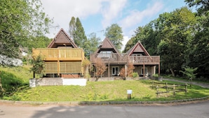 A row of three wooden cabins with balconies, set in a grassy area with trees in the background. - 42 Valley Lodge (Gunnislake)