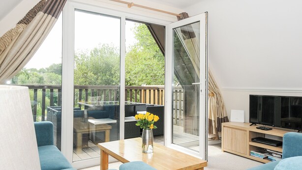A modern living room with a large sliding glass door leading to an outdoor deck, a wooden coffee table with a vase of flowers, and a television on a wooden stand. - 42 Valley Lodge (Gunnislake)