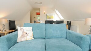 A teal sofa with a cushion featuring a rabbit and the text "Think happy thoughts!" in a room with a sloped ceiling, a dining table, and chairs. - 42 Valley Lodge (Gunnislake)