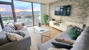 A modern living room with a large window, a flat-screen TV mounted on a brick wall, and a sofa set. - Stunning Penthouse with Balcony | Sunset Views - Pass the Keys (Belfast)
