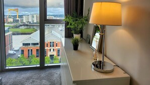 A hotel room with a large window offering a view of a cityscape, a desk with a lamp, and a small plant. - Stunning Penthouse with Balcony | Sunset Views - Pass the Keys (Belfast)